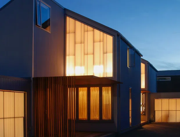 Twilight exterior view of a contemporary multi-storey residential development with sharply articulated volumes, dark vertical cladding, large translucent illuminated windows, timber screening, and integrated garage doors. The buildings face a paved shared driveway or narrow courtyard-like access area, creating a dramatic contrast between the dark facade and warm interior glow.