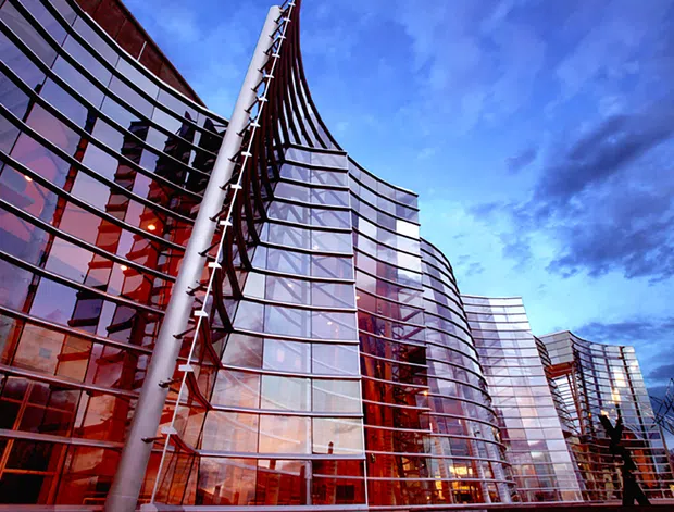 Exterior architectural photograph of a large contemporary commercial building with sweeping curved glass curtain walls, reflective glazing, red-toned interior or facade accents, and prominent exposed structural elements. The building rises against a dramatic blue evening sky and appears to be part of a multi-building urban development.