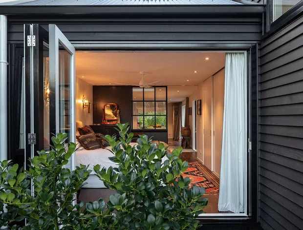 View from outside through a large open glazed doorway into a warmly lit contemporary living room. The dark horizontal exterior cladding frames the opening, while inside are a sofa, rug, artwork, pendant and recessed lighting, curtains, and a glazed internal partition. Dense foreground planting partially obscures the lower facade.
