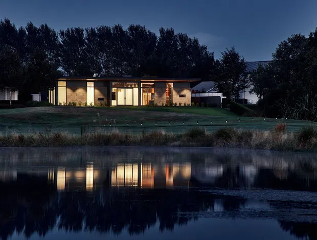 A low, single-storey modern house viewed across a calm pond at dusk. The residence has a long horizontal form, extensive illuminated glazing, warm interior lighting, dark rooflines, and landscaped grounds with ornamental grasses. The building and surrounding trees are reflected in the water, creating a quiet, atmospheric exterior scene.