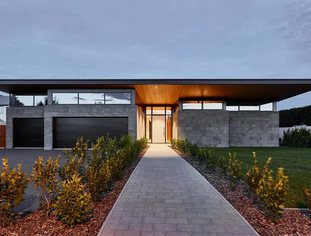 A contemporary single-storey house viewed from the front, with a broad flat roof, deep cantilevered eaves, dark-framed glazing, exposed concrete walls, timber-lined entry soffit, and integrated double garage doors. A straight paved walkway leads through landscaped planting beds to the recessed entrance.