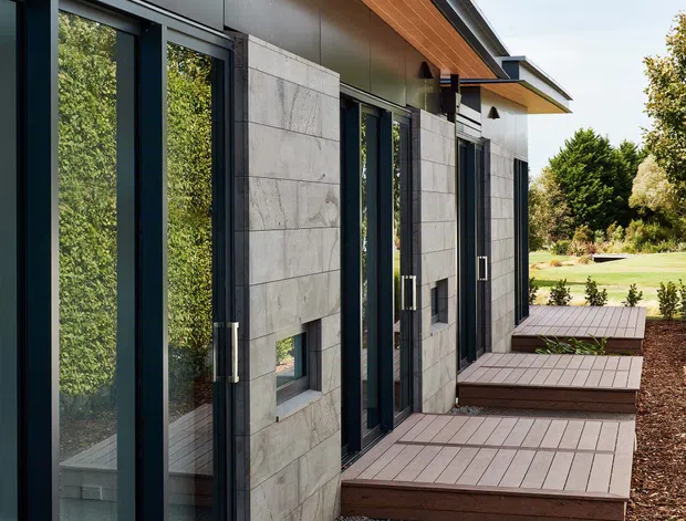 Exterior view of a contemporary single-storey building with large dark-framed glazed doors and windows, grey stone or concrete wall panels, a projecting timber-lined roof soffit, and stepped timber decking platforms. The building opens onto a landscaped garden with mature planting.