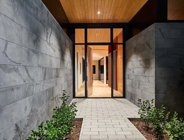 A landscaped entrance walkway leads between tall grey concrete block walls toward a glazed front entry. The path is paved with light rectangular slabs and bordered by low green shrubs and mulch beds. A deep timber-lined soffit extends over the approach, with black-framed full-height glazing and a warm timber-lined interior visible beyond. The composition is modern, geometric, and strongly defined by the contrast between timber, dark frames, and concrete masonry.