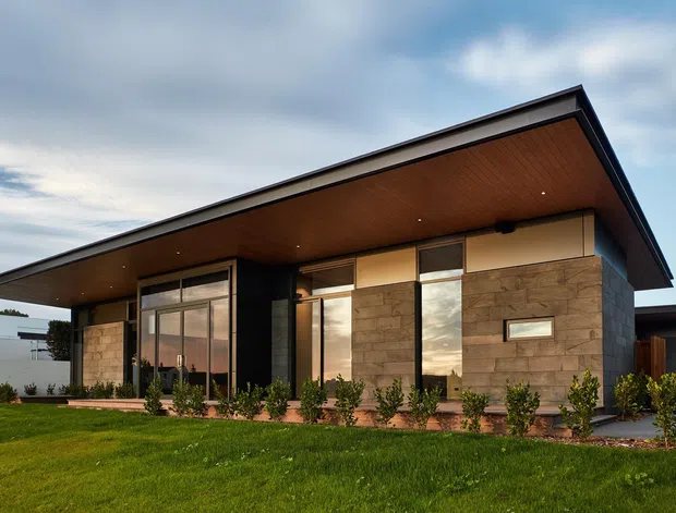Single-storey contemporary house viewed from the front across a lawn. The building has a broad low-pitched roof with a deep projecting eave, warm timber soffit, dark trim, extensive floor-to-ceiling glazing, dark vertical cladding, and stone-clad wall sections. Low landscaped planting lines the facade.