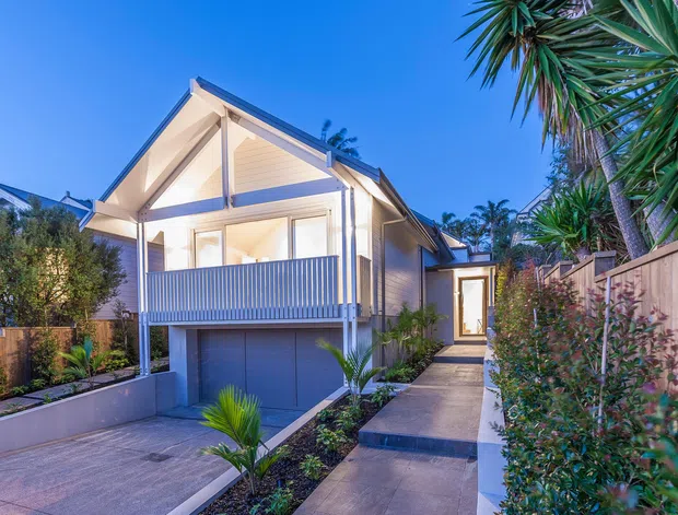 A dusk exterior view of a compact raised home with a prominent front gable, light weatherboard cladding, white trim, and a covered upper-level balcony with vertical balustrade. The lower level contains a garage, while a paved driveway and walkway lead through landscaped planting toward the side entrance. Timber fencing and tropical-style plants frame the property.