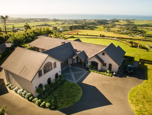 Aerial view of a large light-coloured rural residence with multiple pitched, gabled roof sections arranged around a central courtyard and driveway. The property sits amid expansive green farmland with distant coastal water visible on the horizon.