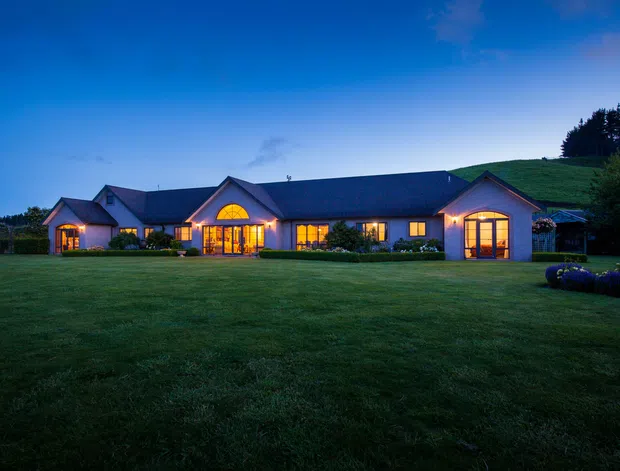 A wide twilight exterior view of a long, low-slung residential building set behind an expansive lawn. The house has a dark pitched roof, pale rendered walls, multiple projecting gabled wings, and warmly illuminated arched-top windows and glazed doors. Dense trees and landscaped planting frame the property, creating a rural estate-like setting.