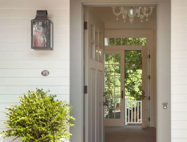 A bright residential entrance viewed from the exterior, featuring pale horizontal weatherboard cladding, a paneled front door within a deep trimmed surround, a wall-mounted lantern, house number plaque, and a large planter. The open entry leads through a tiled hallway toward glazed rear doors and greenery beyond. Traditional architectural detailing and abundant natural light create a welcoming character.