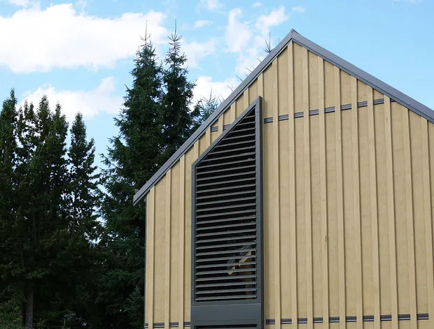 Exterior view of a contemporary gable-ended building clad in vertical pale timber boards, with dark metal edge trims and a tall louvred opening running up the facade. The building sits beside dense trees and a gabion retaining wall under a bright blue sky.