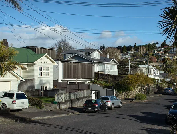 Street-level view of a suburban residential neighbourhood on a sloping site, with detached multi-storey homes, pitched roofs, weatherboard-style facades, balconies, overhead utility wires, paved roadways, and mature vegetation. The image is primarily an exterior streetscape rather than a single interior space.