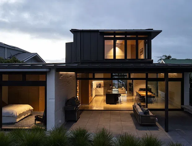 A contemporary two-storey residence photographed at dusk, with extensive black-framed glazing opening onto a paved outdoor living area. The lower level features warm timber-toned interiors, a visible bedroom at left, an open living area, and a freestanding white masonry fireplace at right. A dark-clad upper pavilion with illuminated windows projects above the broad, low roofline.