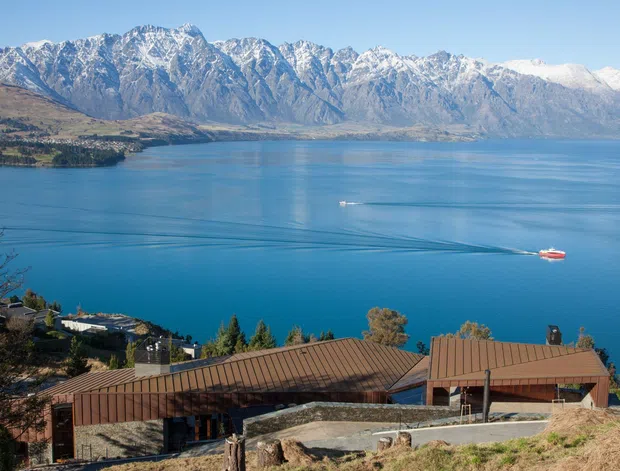 A wide elevated exterior view of a low-rise building with brown metal pitched roofs in the foreground, overlooking a vivid blue lake with boats and dramatic snow-capped mountain ranges beyond. The scene appears to be a mountainous lakeside setting with a clear, bright atmosphere.