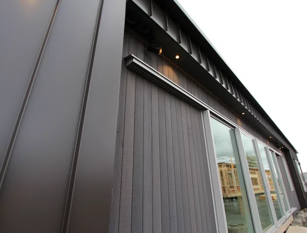 Close-up exterior view of a contemporary dark-clad building, featuring vertically ribbed wall cladding, standing-seam metal panels, deep roof-edge detailing with recessed downlights, and tall glazed openings. The image is taken from outside at a low angle, with a gravel-covered site and a partial interior visible through the windows.