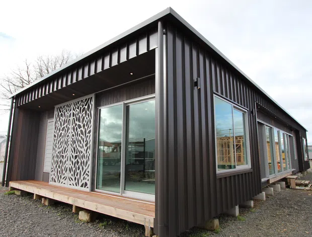 Single-storey contemporary modular-style building with dark vertically ribbed metal cladding, a projecting flat roof canopy, extensive glazing, and a timber deck. A decorative white perforated screen frames the sliding glass entry, while the building is raised slightly above a gravel site on concrete supports.