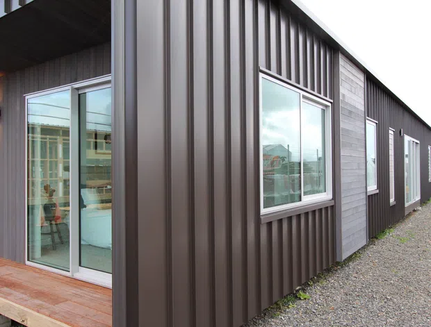Exterior view of a contemporary single-storey building with dark vertically ribbed metal cladding, contrasting grey weatherboard panels, aluminium-framed windows and glazed sliding doors. A timber deck fronts the glazed opening, with gravel surrounding the structure.