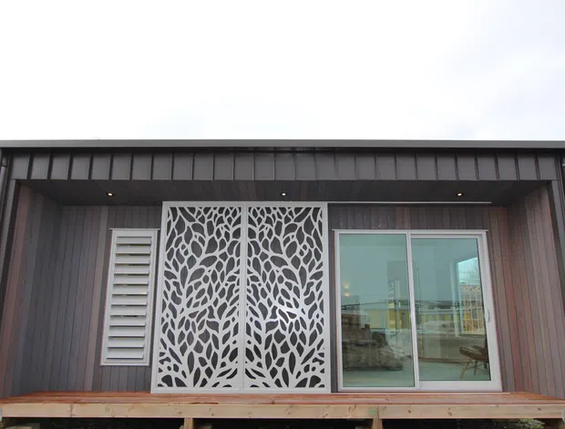 Exterior view of a compact contemporary building with dark vertical cladding, a shallow skillion-style roof with broad eaves, a raised timber deck, large sliding glazed doors, a louvred ventilation window, and a decorative white cut-out screen panel.