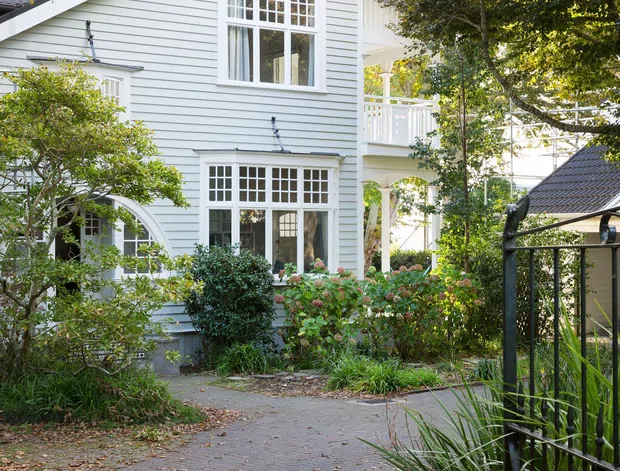 A bright exterior view of a traditional white weatherboard house set within a leafy garden. The two-storey facade features a steep front gable, dark tiled pitched roofing, white multi-pane windows, decorative gable detailing, and a small upper-level balcony. A paved garden path and black metal gate lead toward the house, with mature planting framing the scene.