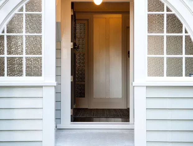 Front entrance of a traditional residential house, featuring pale blue-grey horizontal weatherboard cladding, a white arched entry surround, paired arched textured-glass sidelights, and a warm timber door. Patterned black-and-white tiled stair risers lead up to the recessed entry, which is lit by a hanging pendant.