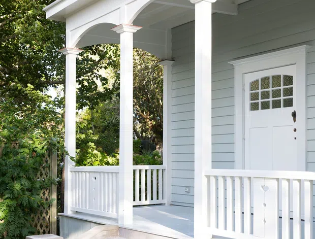 Bright exterior view of a traditional residential front porch with white timber columns, decorative arches, balustrades, and grey-blue weatherboard cladding. The raised entry is accessed by concrete steps and includes a white glazed front door, garden planting, and blue ceramic planters.