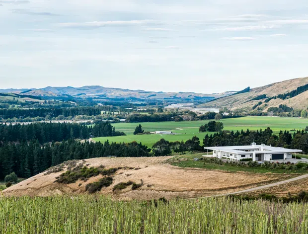 A wide rural landscape view overlooking a modern, low-profile white house set among green fields, rows of vegetation, wooded areas, and rolling hills. The property is reached by a curving driveway and sits prominently within an expansive agricultural setting beneath a bright, lightly clouded sky.