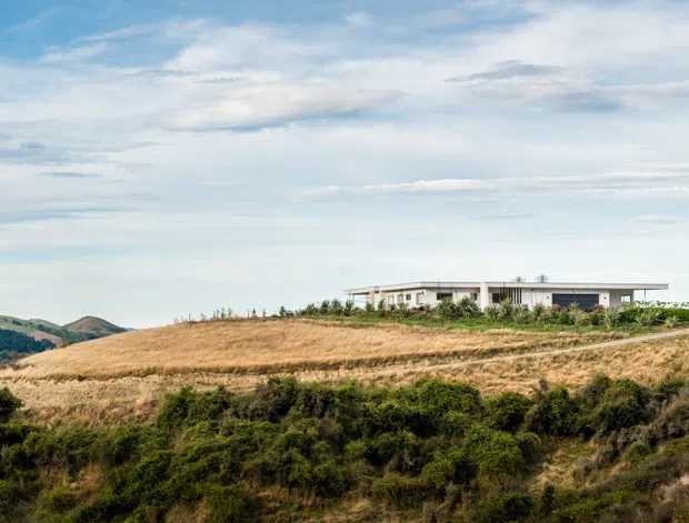 A low, elongated modern house set on a gently elevated rural or coastal hillside, surrounded by dry grass and low scrub. The building has a flat roof, extensive horizontal glazing, pale exterior walls, and a restrained minimalist form. Rolling hills and a broad cloudy sky form the distant backdrop.