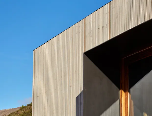 Close-up exterior view of a modern building facade with vertically ribbed light timber cladding, a deep dark recessed entry opening, and a timber-framed door. Strong sunlight creates sharp geometric shadows against the facade, with clear blue sky and a low natural hillside in the background.
