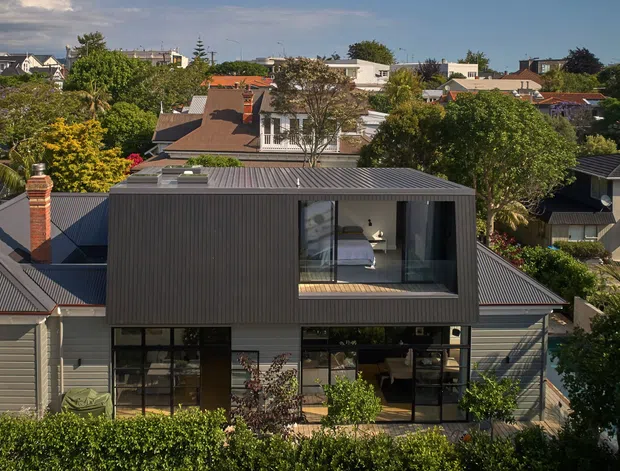 Street-facing view of a contemporary residential extension in a leafy suburban neighbourhood. The dark vertically clad upper volume projects over a glazed ground floor, with a low-pitched metal roof, large black-framed windows and doors, and a recessed upper-level bedroom or living space opening onto a balcony. Established gardens and surrounding pitched-roof homes are visible in the background.