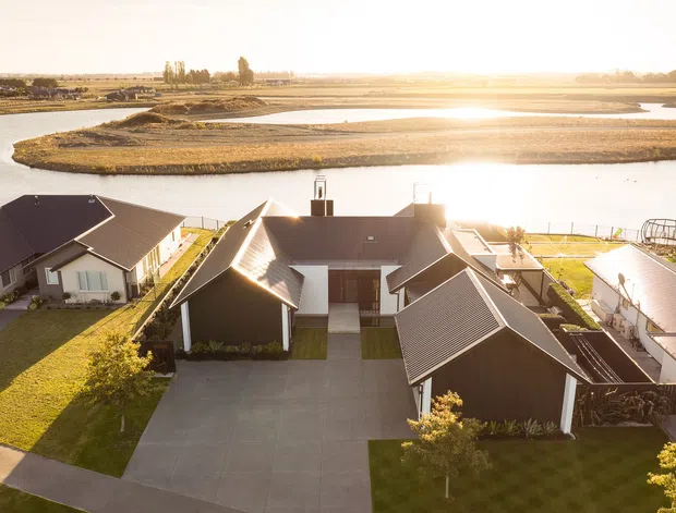 Aerial view of a contemporary low-rise residential development beside a broad tidal or wetland waterway. Several detached dark-roofed homes are arranged around paved driveways and manicured lawns, with the central house featuring multiple gabled roof forms, dark vertical cladding, and light-coloured garage and wall elements. The sunlit water and landscaped peninsula form the background.