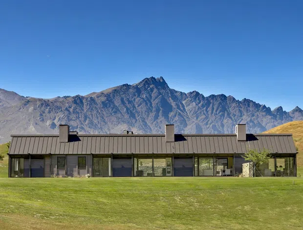 A long, low single-storey contemporary house set in an expansive open grassy landscape, with a dark standing-seam pitched roof, extensive front glazing, and rugged mountain ranges directly behind it. The building has a horizontal composition and restrained dark-and-neutral material palette, with a few planted trees and rolling dry hills around the site.