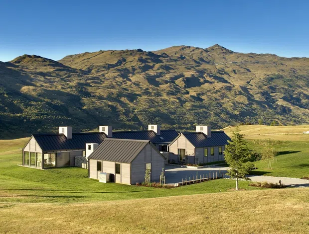A wide exterior view of a contemporary rural residential development set in an expansive grassy valley, with several dark-gabled buildings clustered in the foreground and a rugged mountain range rising behind them. The buildings feature pitched roofs, dark metal roofing, light cladding, large glazed openings, and a restrained modern country aesthetic.