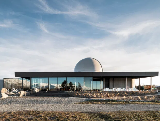 A low, single-storey modern building with an expansive flat roof, extensive floor-to-ceiling glazing, dark horizontal cladding, and a prominent curved dome-like roof element. The structure sits on a sparsely landscaped gravel site with exposed rock, under a bright open sky. A neighbouring house is visible at the far left.
