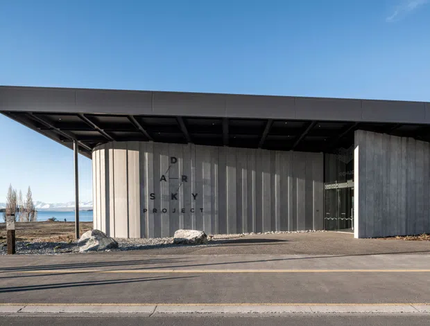 Exterior view of a low-profile contemporary building with a broad flat roof and deep cantilevered overhang. The facade combines dark metal cladding with vertical grey concrete or timber-like paneling, and the entrance is sheltered beneath the roof. A lake and mountains are visible in the background, though the building is the primary subject.