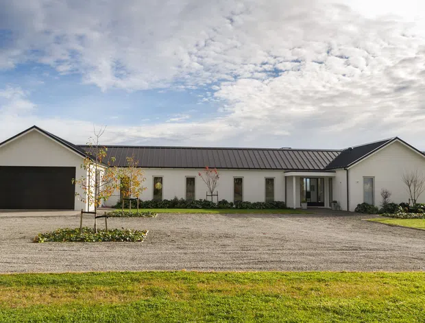 Single-storey contemporary residential building viewed from the front, with a long low-profile form, pale rendered walls, dark standing-seam pitched roof, attached double garage, narrow vertical windows, central glazed entry, gravel driveway, and landscaped lawn with young trees.