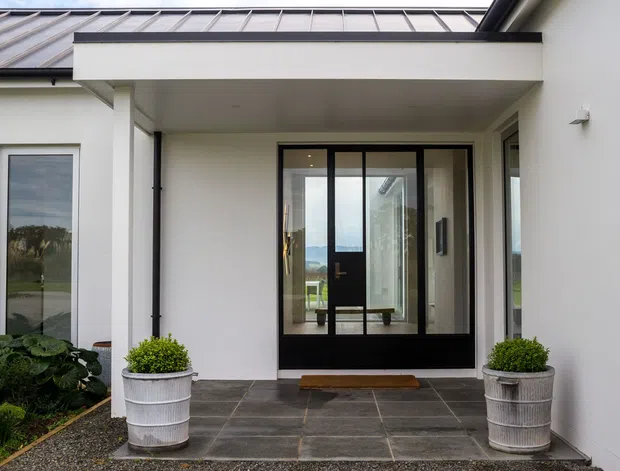 Front entrance of a contemporary home, featuring a covered porch, black-framed glazed entry door, white rendered walls, dark tiled paving, and a low-profile metal roof. Symmetrical potted greenery frames the doorway, with distant landscape visible through the glazing.