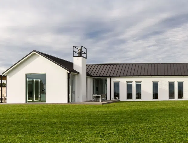 A wide exterior view of a low, single-storey white residence set behind an expansive lawn. The home has a long linear floor plan with several pitched gable-roofed wings connected by a central section with a dark metal roof, tall glazed doors and windows, a covered outdoor area, and a simple chimney. The setting appears open and suburban or rural, with a restrained contemporary farmhouse-like appearance.