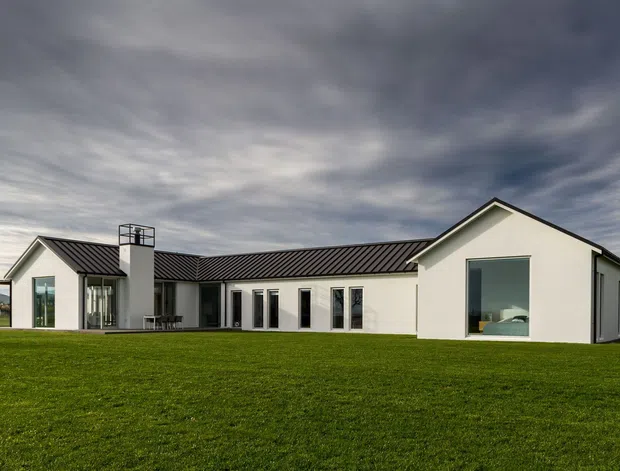 A low, single-storey contemporary house set behind an expansive lawn. The white rendered facade has strong gabled volumes, a dark standing-seam pitched roof, tall narrow windows and glazed doors, with a prominent chimney and a simple linear connecting wing. The image is an exterior architectural view under a dramatic overcast sky.