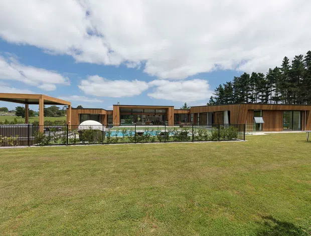 Wide exterior view of a low, single-storey contemporary residence set in a broad landscaped lawn. The building has long horizontal timber-clad wings, extensive dark-framed glazing, and a central courtyard-like pool area enclosed by a black metal fence. A simple timber pergola stands to the left, with mature trees behind the house and open rural landscape beyond.