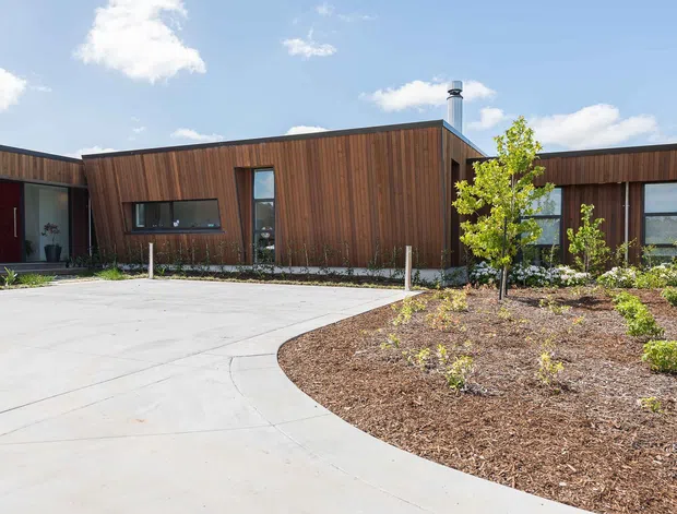 Single-storey contemporary building with a low, angular roofline and extensive vertical timber cladding. Dark-framed windows and glazed doors punctuate the facade, with a curved concrete driveway and newly landscaped garden beds in the foreground. The image is photographed from outside under a bright blue sky.