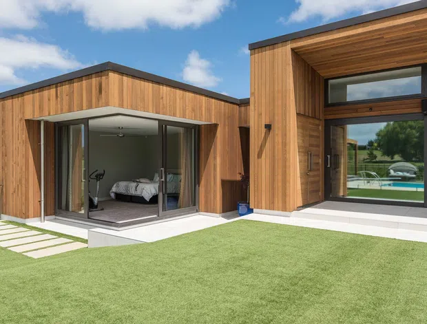 Contemporary single-storey residence with two intersecting timber-clad volumes, dark flat rooflines, extensive floor-to-ceiling glazing, and a sheltered recessed entry. The left glazed room reveals a bedroom and exercise equipment, while the larger opening reflects or overlooks a pool and rural landscape. A manicured lawn and stepping-stone path occupy the foreground.