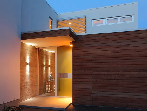 Contemporary residential facade at dusk, featuring a recessed illuminated entryway, extensive horizontal dark timber cladding, smooth pale rendered walls, narrow clerestory windows, and a slatted privacy screen above the entrance.