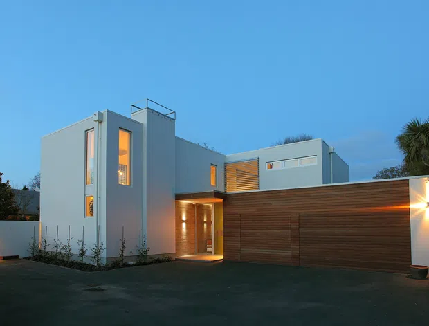 Twilight exterior photograph of a contemporary multi-level residence with crisp white rendered cubic forms, warm timber-clad garage and entry elements, narrow vertical windows, recessed lighting, and a broad paved driveway. Tropical planting and perimeter walls frame the modern facade.