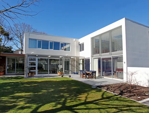 A contemporary white two-storey residence viewed from the landscaped rear garden. The house features a low-slung interconnected form, extensive glazing, a projecting upper volume, large horizontal external blinds, a covered outdoor entertaining area, and a paved terrace opening onto a lawn.