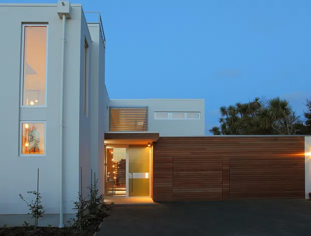 Contemporary two-storey residential facade photographed at dusk. The building has a crisp white rendered exterior, tall vertical glazing, a recessed warmly lit entry, and a broad horizontal timber garage door or screen. Minimal landscaping and spiky ornamental trees frame the driveway, while integrated exterior lighting creates a warm contrast against the blue evening sky.