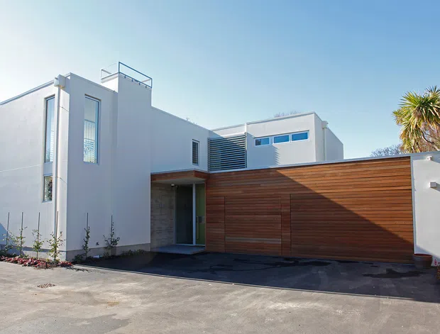 Exterior view of a contemporary multi-level residence with clean white rendered walls, a broad horizontal timber garage door, narrow vertical and clerestory windows, and a flat-roofed composition. The building fronts a paved driveway and includes a small rooftop terrace or balustrade detail.