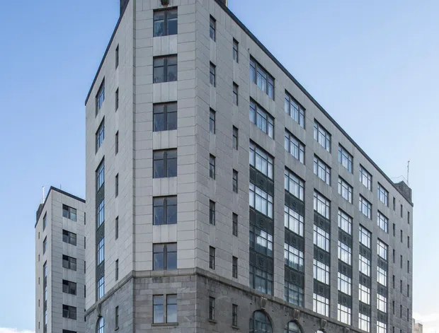 Street-level exterior photograph of a substantial multi-storey urban commercial building. The building has a pale grey stone or concrete-panel facade, a darker stone base, tall repetitive industrial-style windows, large arched windows at ground level, rooftop railings, and a flagpole. It occupies a prominent city corner with adjoining buildings and a broad paved street visible around it.