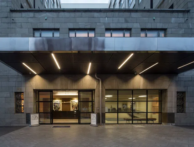 Street-level view of a contemporary commercial building entrance beneath a broad metal-clad canopy. The facade is composed of dark grey stone or concrete masonry, with extensive black-framed glazed entry doors and sidelights. Linear recessed lighting illuminates the sheltered entrance, and a paved forecourt leads into the building. Interior reception or lobby areas are visible through the glazing.