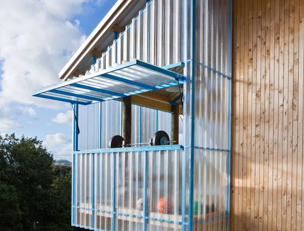 Exterior architectural photograph of a compact elevated structure attached to a timber-clad building. The extension is enclosed with translucent corrugated panels framed in bright blue, with a projecting translucent canopy and visible stored or functional contents behind the panels. Timber posts support the structure above a landscaped sloping site, while the main building has vertical natural timber cladding and exposed timber roof framing beneath a shallow pitched roof. Bright blue sky and vegetation surround the building.