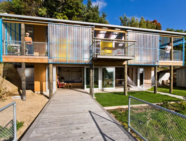Exterior view of a contemporary elevated multi-level residential building set into a sloping, landscaped site. The structure features a long, low flat roof, exposed timber posts, extensive glazing, timber decking and walkways, and blue-grey corrugated or slatted screening around upper-level balconies. Open ground-level living areas are visible beneath the elevated upper floor, with chain-link fencing and lawn in the foreground.