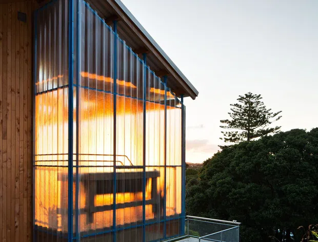 Exterior architectural photograph of a compact elevated timber structure in a landscaped setting. The building has a steeply pitched asymmetrical roof with exposed timber rafters, timber wall cladding, and a prominent translucent corrugated or ribbed facade framed in blue-toned metal. Warm interior light glows through the translucent panels. A raised timber deck and metal mesh balustrade extend from the building, supported on slender posts, with dense vegetation surrounding the site.