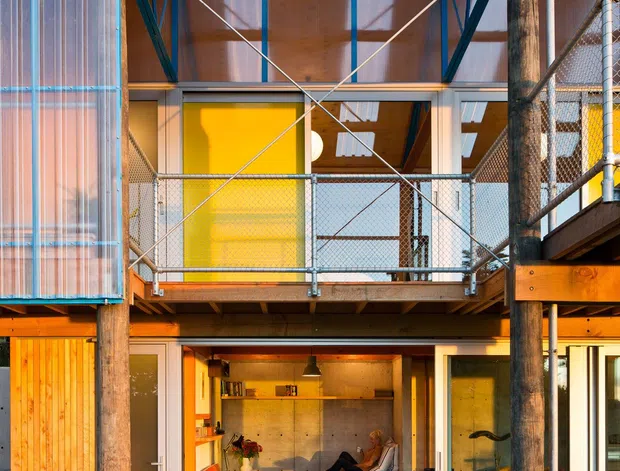 A two-storey residential building viewed from an exterior timber deck and walkway. The facade combines warm timber cladding and exposed timber posts with steel bracing, chain-link balcony railings, translucent corrugated panels, and extensive glazing. An open upper-level balcony with a bright yellow door is visible above a ground-floor living room, which contains a sofa, red armchair, coffee table, and built-in concrete-toned surfaces. The composition highlights exposed structure, indoor-outdoor connection, and a warm industrial-contemporary aesthetic.